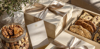 generous selection of luxury gifting items displayed on a polished marble or stone table under cinematic golden hour light. the scene features multiple artisan confections, cookies, crackers, nuts.