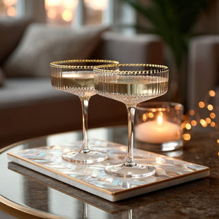 Two elegant coupe glasses with gold-rimmed edges filled with champagne, placed on a glossy pearl-inlay tray atop a glass coffee table, with a lit candle and soft bokeh lights creating a warm, festive ambiance in the background.