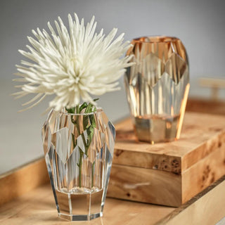 Clear faceted crystal bud vase holding a single white chrysanthemum on a wood shelf, with an amber glass vase softly blurred behind.