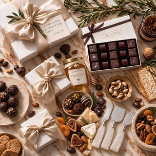 A styled photo of biggest little baskets $750 custom curated gift box featuring an assortment of gourmet chocolates, nuts, cookies, and spreads arranged on an acacia wooden board with two cream gift boxes tied by large dark brown ribbons, on a beige fabric background with alabaster cheese knives and a travertine bowl and tavertine tray. 