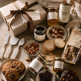 A Styled photo of Biggest Little Baskets $450 Gifting Tier custom curated gift box gourmet gift box of chocolates and spreads from Valerie, displayed in a beautifully styled flat-lay arrangement with cookies, jars of jam, crackers, and nuts on a neutral fabric background.
