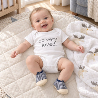 Styled photo of the Baby Boy Essentials gift Box showing a baby boy wearing the so very loved onesie and striped blue and white vegan leather booties with the white with moose design muslin throw blanket/swaddle from Biggest Little Baskets