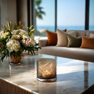 Brown cut glass hurricane container with a lit votive candle, styled on a marble coffee table in a sunlit Malibu-inspired living room with luxury throw pillows, soft floral arrangement, large windows, and ocean-view backdrop.