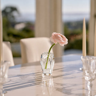 A single pale pink flower in a small clear faceted glass vase set on a polished dining table, with chairs and a softly blurred outdoor background.