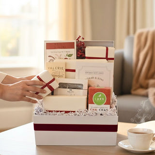 Hands lifting a small ribbon-wrapped treat from the open Crimson & Cream holiday gift box styled beside a teacup in a softly lit living room.