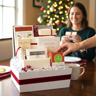 Woman reaching into an open Crimson & Cream holiday gift box filled with cookies, chocolate, nuts, caramels, and a ceramic ornament beside a Christmas tree.