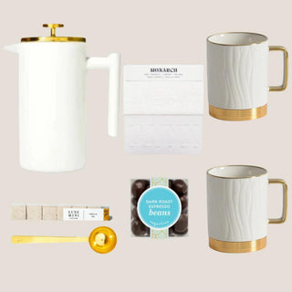Deluxe Java Joy contents arranged on a neutral background: French press, two mugs, whole-bean coffee, vanilla sugar cubes, espresso beans, and gold scoop.
