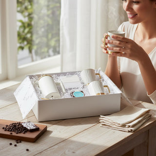 Woman holding a porcelain mug beside an open Deluxe Java Joy coffee gift box with white-and-gold French press, coffee, mugs, sugar cubes, scoop, and espresso beans.