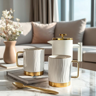 Matte white and gold French press with two cream porcelain mugs and gold scoop styled on a marble coffee table in a modern living room.