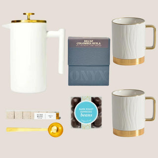 Deluxe Java Joy decaf option shown with white-and-gold French press, two porcelain mugs, Colombia Huila coffee, vanilla sugar cubes, espresso beans, and scoop.