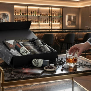 Open black Deluxe Sip & Savor whiskey gift box styled on a modern bar counter beside a hand holding a clear whiskey tumbler.