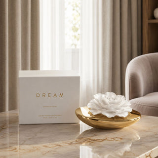 Dream white porcelain flower diffuser in a gleaming gold ceramic vessel beside its presentation box on a marble table in a softly lit room.