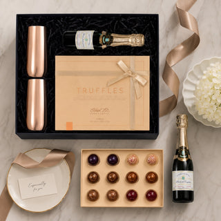 Luxury champagne and truffle gift set arranged on a light marble surface. The set includes a black gift box with two rose gold champagne flutes, a mini unscented champagne bottle candle, a beige Truffles box, a tray of assorted chocolates, a small plate with a card, and a gold ribbon.