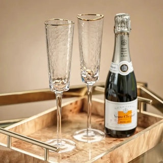 Two gold-rim hammered champagne flutes standing beside a bottle of sparkling wine on a warm metallic serving tray.
