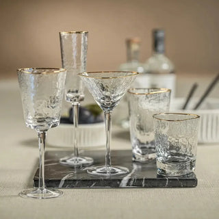 Gold-rim hammered martini glass centered among coordinating champagne, wine, highball, and tumbler glassware on a dark serving board.