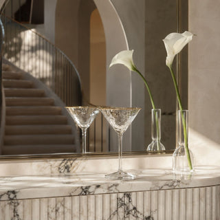 Two gold-rim hammered martini glasses styled on a veined marble console beneath an arched mirror with white calla lilies.