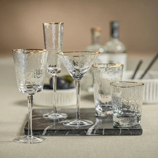 Gold-rim hammered wine glass displayed with coordinating champagne, martini, highball, and tumbler glassware on a dark serving board.