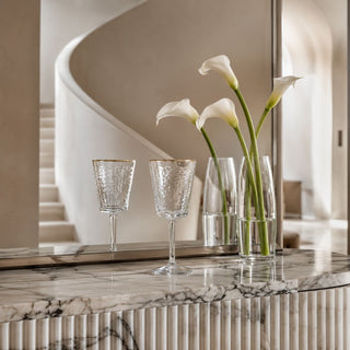 Two gold-rim hammered wine glasses styled on a veined marble console beneath an arched mirror with three white calla lilies.