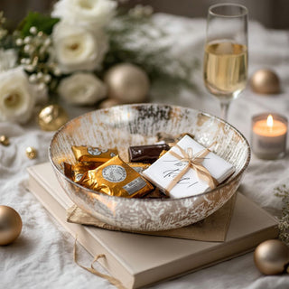 Gold and silver foiled glass bowl filled with wrapped chocolates and styled on white books at a candlelit holiday table.