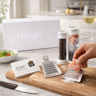 Luxury Rivsalt gourmet salt tasting set displayed on a light stone kitchen countertop, featuring a white Rivsalt gift box, stainless steel salt grater, wooden base, pepper taster, and Himalayan salt rock as a hand grates salt while preparing food in an elegant, modern kitchen setting
