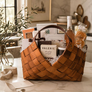 Grand Epicurean large gourmet gift basket on a marble table with woven wood basket, leather handles, alcohol-free sparkling bottle, seasonal sweet and savory foods, and white faux botanicals.