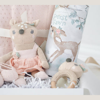 Close view inside the Heirloom Baby Girl gift box showing the knit fawn toy, pink blanket, crocheted booties, fawn ring rattle, and Forest Fawn story cylinder.