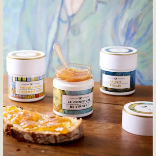Jars of confiture with a spoon on a wooden surface, with a colorful abstract painting in the background.