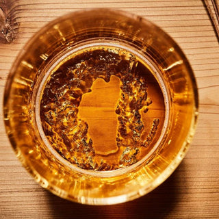Overhead view through amber beer showing the Lake Tahoe-shaped dimensional relief molded into the base of a pint glass on a light wood table.