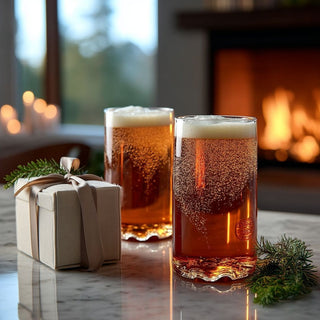 Two Lake Tahoe topographic pint glasses filled with amber beer on a marble table beside a wrapped gift and evergreen sprigs, with a fireplace behind them.