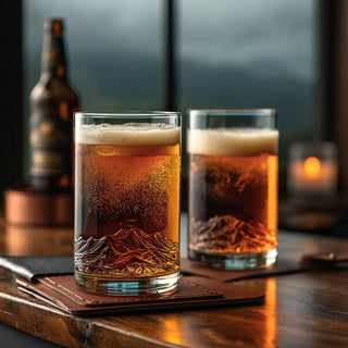 Two Lake Tahoe topographic pint glasses filled with golden beer on a dark home bar, with the molded mountain relief visible in the foreground glass.