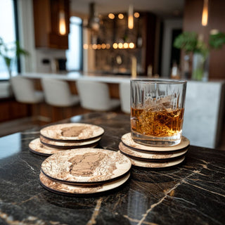 Lake Tahoe topographic wood coasters arranged on a dark marble home bar, with an amber whiskey glass resting on one etched birch coaster.