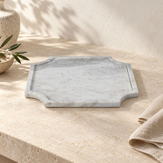 The 14-inch Volakas marble tray displayed on a beige stone surface beside ceramics, greenery, and folded linen, showing its shape and marble veining in natural light.