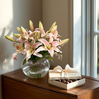 Light pink Casablanca lily faux floral arrangement in a clear glass vase styled on a refined entryway table beside a cream ribbed box of chocolate truffles with satin bow, capturing an elevated quiet luxury floral gifting moment.