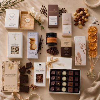 Luxury gourmet gift box flat lay featuring Valerie artisan chocolates, gourmet cookies, premium chocolate bars, candied almonds, chocolate pretzels, dried orange slices, sparkling wine gummies, artisan diffuser, and curated gift box essentials arranged on a warm limestone surface. Editorial-style luxury gift basket photography by Biggest Little Baskets for concierge gifting, corporate gifts, client appreciation, holiday gifts, and luxury gift delivery.