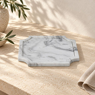 Medium Volakas marble tray on a beige stone surface beside ceramic vessels, leafy branches, and folded linen, showing its gray veining and shaped edge in sunlight.