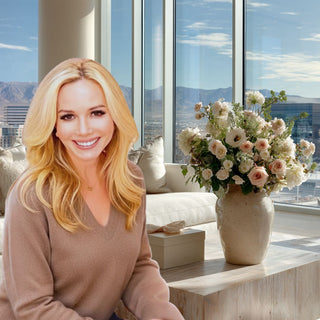 The founder of Biggest Little Baskets Mel is sitting in a modern living room with large windows over looking reno nevada. There is a vase of flowers on a table & a gift box wrapped beautifully next to her.