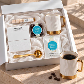A curated coffee gift set in a white box, including Monarch coffee boxes, dark roast whole bean pods, a gold spoon, and two white mugs with gold bases, arranged on a stone surface with coffee and beans nearby.