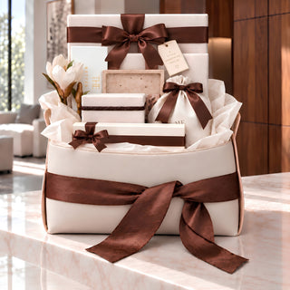 A luxury gift basket arrangement featuring cream-colored boxes and a white drawstring pouch, all elegantly tied with wide brown satin ribbons and bows. The set is styled with white tulips and tissue paper on a polished marble countertop in a modern, sunlit interior.