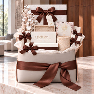 A curated gift basket set on a marble countertop in a bright, modern room. The set includes a cream-colored gift box with a large brown satin ribbon, a candle, a boxed item labeled 'THE DATE SPOT', dried floral branches, and other matching cream and brown gift items.