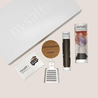 RIVSALT seasoning gift set arranged beside its matte white box with stainless steel grater, round wooden stand, salt rock tasters, Java long pepper, and conversation cards.