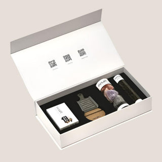 Open matte white RIVSALT gift box showing the grater, wooden stand, salt rock tasters, Java long pepper, and conversation cards secured in a fitted black insert.