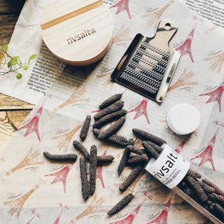 RIVSALT Java long pepper pieces arranged beside the stainless steel grater, wooden stand, and pepper taster tube on printed paper.