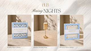 A styled product collage under the heading 'BLB Rainy Nights' on a light marble-textured background. Three vertical panels show coordinated items: a light blue Scrabble game box, a small gold wireless USB mini lamp, and another light blue Monopoly game box.