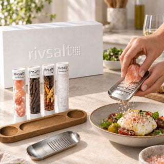 Hands grate Himalayan salt over a tomato and burrata salad on a kitchen countertop, with the Rivsalt gift box, wooden stand, and seasoning tubes nearby. A serving idea for home cooks and hosts.
