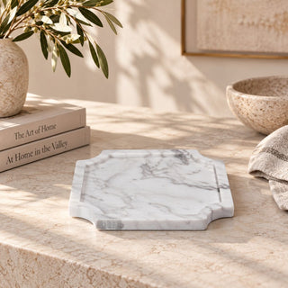 Small Volakas marble tray displayed on a sunlit stone tabletop beside stacked books, a leafy arrangement, a ceramic bowl, and folded fabric.