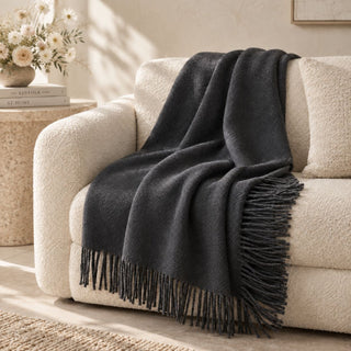 Stargazer charcoal throw with dark fringe draped over a cream bouclé sofa beside a stone table and flowers.