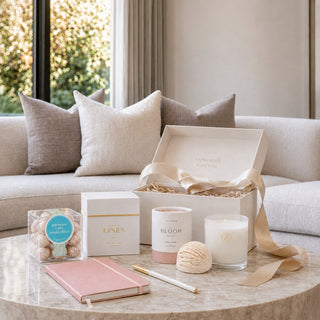 A curated gift set arranged on a round marble table in front of a light sofa. The set includes an open cream gift box with a beige ribbon, a pink notebook, a pink box labeled LINEN, a candle jar labeled BLOOM, a clear container of bath bombs, a bath bomb, and a gold pen. Birthday Wishes Gift Box styled in a modern living room with journal, candle, bath truffle, pen, and cookie bites