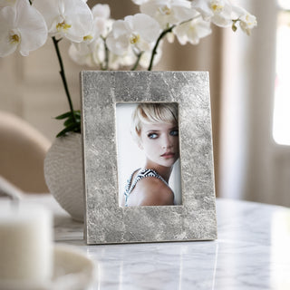 A square silver picture frame with a textured metallic finish, holding a portrait photo of a woman, displayed on a marble table with white orchids in the background.