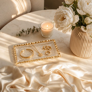 A styled flat-lay of gold jewelry, including a cuff bracelet, a necklace with a heart pendant, and two rings, arranged on a clear glass tray with a gold beaded border. The tray sits on a marble tablecloth next to a lit candle, white flowers in a ribbed vase, and draped champagne silk.