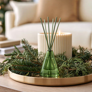 A green glass reed diffuser with several green reed sticks stands on a round gold tray, surrounded by pine branches in front of a cream ribbed candle in a softly blurred living room setting.